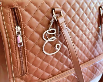 May include: A tan quilted handbag featuring a silver-toned metal letter E charm attached to the strap hardware. The bag includes a vertical zippered pocket with a silver pull, matching tan stitching, and two vertical straps attached to the top.