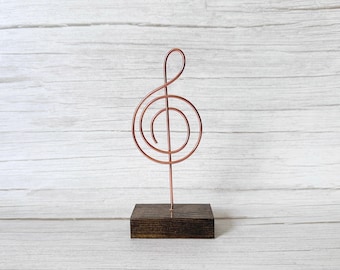 May include: A copper-colored metal wire sculpture shaped like a treble clef musical symbol. The wire form is mounted vertically into a rectangular, dark brown stained wooden base with a visible wood grain texture throughout the piece.