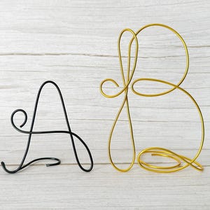 May include: A black wire letter A and a gold wire letter B are displayed against a light wood grain background. The letters are stylized with a cursive font. The letter A is black, and the letter B is gold. The letters are decorative.