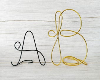 May include: A black wire letter A and a gold wire letter B are displayed against a light wood grain background. The letters are stylized with a cursive font. The letter A is black, and the letter B is gold. The letters are decorative.