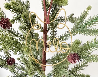 May include: A gold-colored wire ornament shaped like a cat head with the name "milo" written in cursive script inside the circular frame. The ornament is suspended by a thin, dark red cord and features a continuous wire design.