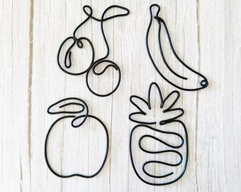 May include: Black wire fruit sculptures of a cherry, banana, apple, and pineapple. The minimalist designs are placed on a light wood-grain surface. The cherry has two cherries and a stem. The banana is curved. The apple has a stem and a leaf. The pineapple has a spiky top.