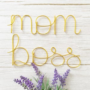 May include: Gold wire sculpture of the words "mom boss" on a white wood background with purple flowers in the bottom right corner.