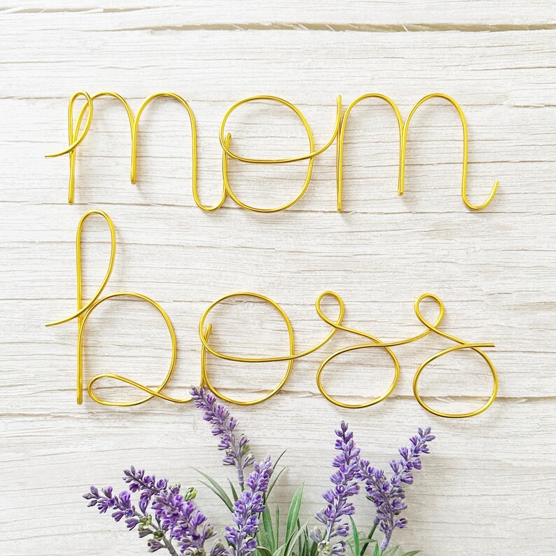 May include: Gold wire sculpture of the words "mom boss" on a white wood background with purple flowers in the bottom right corner.