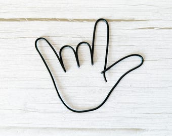 May include: Black metal hand sign sculpture, a decorative piece. The hand is in the 'I love you' gesture, with the thumb, index finger, and pinky extended. The sculpture is set against a white wooden background.