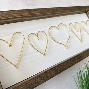 May include: A wooden frame with a white background and five gold wire hearts. The hearts are arranged in a row, with the largest heart on the left and the smallest heart on the right.