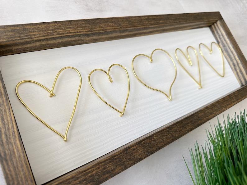 May include: A wooden frame with a white background and five gold wire hearts. The hearts are arranged in a row, with the largest heart on the left and the smallest heart on the right.