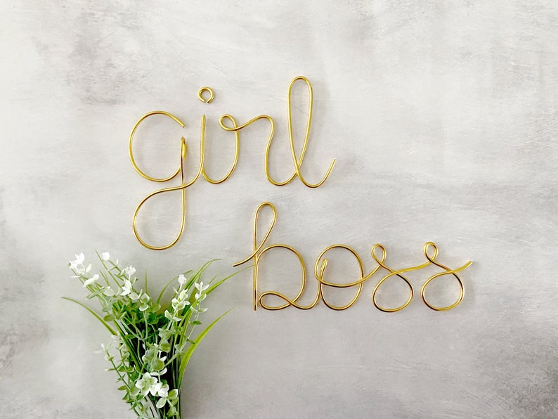 May include: Gold wire sculpture of the words "girl boss" on a white background.