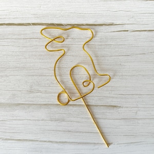 May include: A gold-colored wire bunny cake topper. The bunny is crafted from a single piece of wire, with a looped heart shape at the bottom. The topper is designed to be inserted into a cake or other dessert for decoration.
