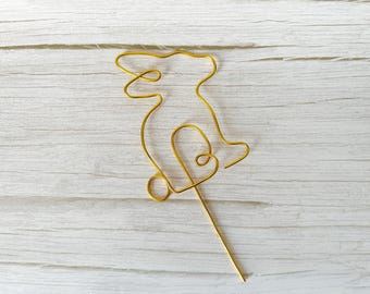 May include: A gold-colored wire bunny cake topper. The bunny is crafted from a single piece of wire, with a looped heart shape at the bottom. The topper is designed to be inserted into a cake or other dessert for decoration.