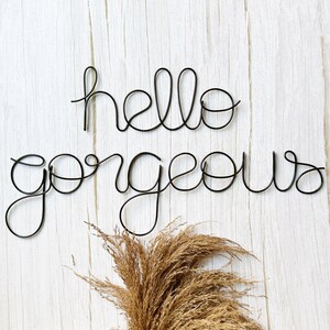 Puede incluir: Arte de pared de alambre negro con la frase "hello gorgeous" sobre un fondo de madera blanca. El texto en cursiva está en una fuente delicada y fluida. Hierba ornamental seca de color marrón claro en la parte inferior de la imagen.