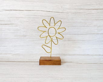 May include: A gold-colored wire sculpture shaped like a flower with eight petals and a single leaf on the stem. The wire flower is mounted vertically into a rectangular, dark brown stained wooden base that provides a stable foundation.