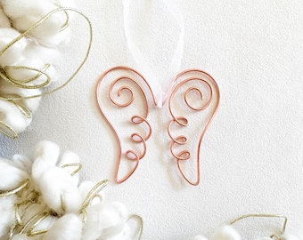 May include: A copper-colored wire ornament shaped like a pair of angel wings, featuring coiled wire details and a thin white ribbon loop at the top for hanging. The ornament is centered against a plain, light-colored background.