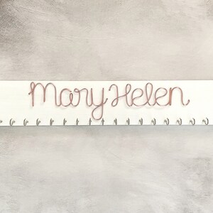 May include: A white wooden sign with the name "Mary Helen" written in copper wire script. Below the name are several small silver hooks. The sign is designed for hanging items.