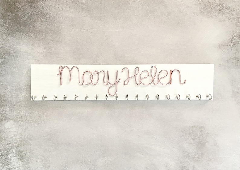 May include: A white wooden sign with the name "Mary Helen" written in copper wire script. Below the name are several small silver hooks. The sign is designed for hanging items.
