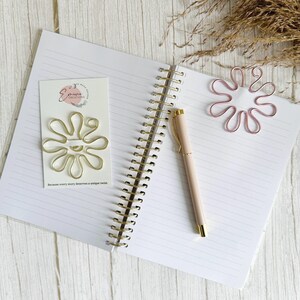 May include: An open notebook with lined pages, a gold spiral binding, and a light pink pen. A gold wire flower-shaped paper clip is on a white card with text. A pink wire flower-shaped paper clip rests on the notebook pages. Dried grass is in the background.