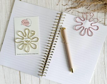 May include: An open notebook with lined pages, a gold spiral binding, and a light pink pen. A gold wire flower-shaped paper clip is on a white card with text. A pink wire flower-shaped paper clip rests on the notebook pages. Dried grass is in the background.