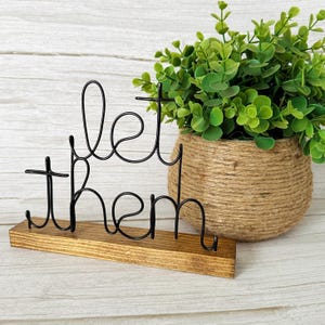 May include: Black wire sculpture of the words "let them" on a wooden base. The sculpture is in front of a potted plant with green leaves.
