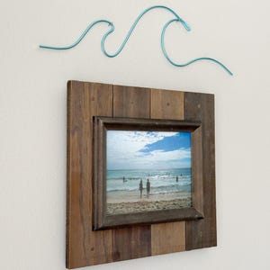 May include: A rustic wooden picture frame with a beach scene photo inside. A blue wire wave sculpture is hanging above the frame.