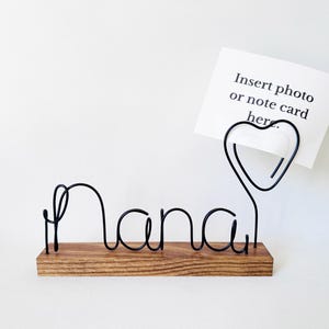 Puede incluir: Un soporte de alambre negro con la palabra Nana en letra cursiva sobre una base rectangular de madera. Un clip de alambre en forma de corazón sostiene una tarjeta blanca con el texto negro Insert photo or note card here.
