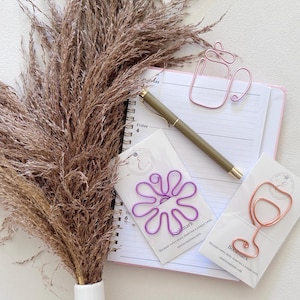 May include: A spiral-bound planner with a gold pen, a pink wire coffee cup paperclip, a purple wire flower paperclip on a white card, and a rose gold wire wine glass paperclip on a white card, accompanied by a large bunch of dried brown pampas grass.