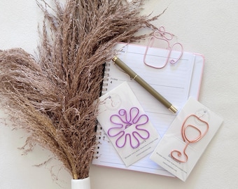 May include: A spiral-bound planner with a gold pen, a pink wire coffee cup paperclip, a purple wire flower paperclip on a white card, and a rose gold wire wine glass paperclip on a white card, accompanied by a large bunch of dried brown pampas grass.