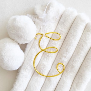 Puede incluir: Un adorno de alambre dorado con la letra E en letra cursiva, sujeto a un calcetín de piel sintética blanca con dos pompones blancos y una cinta blanca fina, creando un accesorio decorativo suave y con textura.