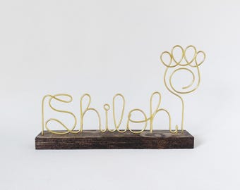 May include: A gold-colored wire sculpture spelling the name Shiloh in cursive script, attached to a rectangular dark brown wooden base. To the right of the name is a wire outline of a paw print with four rounded toes and a central spiral.