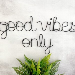 May include: Black wire wall decor with the words "good vibes only" on a white background with a green plant in the bottom center.