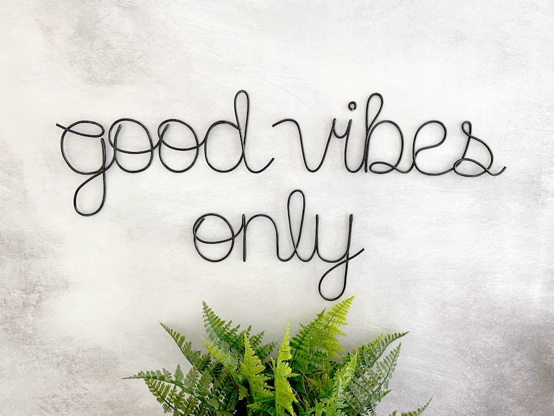 May include: Black wire wall decor with the words "good vibes only" on a white background with a green plant in the bottom center.