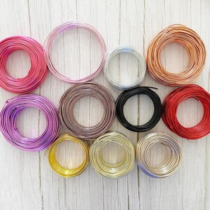 May include: A selection of colourful wire spools in shades of pink, purple, silver, copper, black, and red. The spools are arranged on a light-coloured wooden surface, displaying the vibrant colours and textures of the craft wire.
