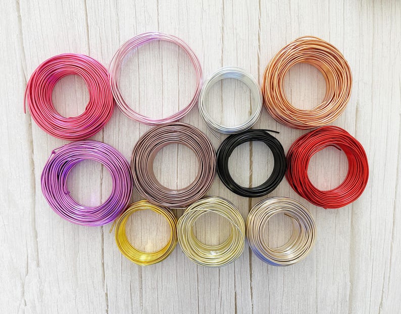 May include: A selection of colourful wire spools in shades of pink, purple, silver, copper, black, and red. The spools are arranged on a light-coloured wooden surface, displaying the vibrant colours and textures of the craft wire.