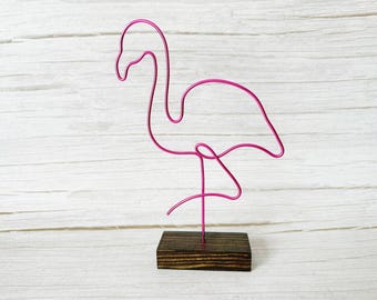 May include: A pink wire flamingo sculpture on a dark stained wooden base. The flamingo is crafted from a single continuous wire, depicting the bird's form in a minimalist style. The sculpture is set against a white wooden background.