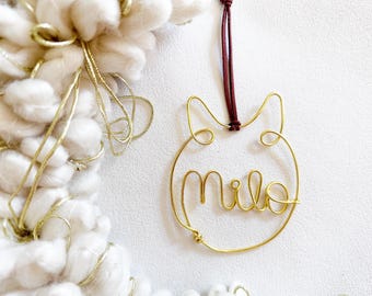 May include: A gold-colored wire ornament shaped like a cat head with the name Milo written in cursive script inside the circular frame. The ornament hangs from a thin dark red cord and features a simple, continuous wire design.