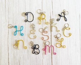 May include: A collection of twelve individual letter keychains crafted from colored wire in various cursive fonts. Each letter features a small metal split ring attachment. The colors include black, gold, yellow, turquoise, pink, and copper tones.