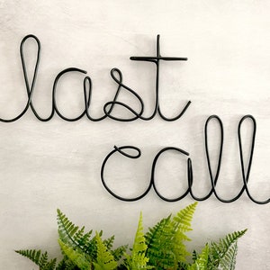 May include: Black metal wire sculpture of the words "last call" hanging on a white wall.