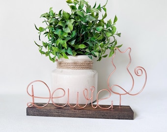 May include: A copper-colored wire sculpture featuring the name "Dewie" in cursive script, connected to a stylized cat silhouette. The wire art is mounted on a rectangular dark brown wooden base, creating a decorative display piece.