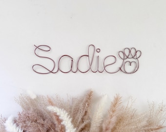 May include: A metallic rose gold wire wall art piece featuring the name Sadie in a continuous cursive script, followed by a stylized paw print icon that incorporates a small heart shape within its central design element.
