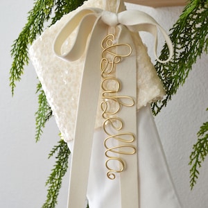 Puede incluir: Un calcetín navideño blanco con un puño de lentejuelas y una cinta larga de color crema atada en un lazo. Sujeta a la cinta hay una etiqueta de alambre dorado con el nombre Isabella escrito en cursiva, creando un adorno festivo.