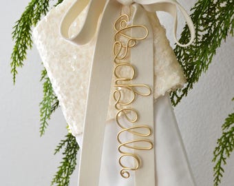 May include: A white Christmas stocking featuring a sequined cuff and a long cream-colored ribbon tied in a bow. Attached to the ribbon is a gold-colored wire name tag shaped into the cursive script name Isabella, creating a decorative holiday accent.