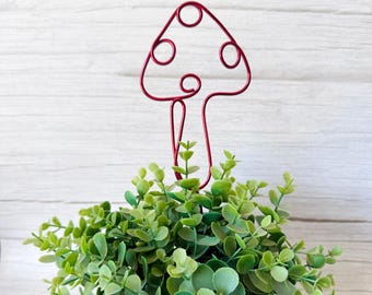 May include: A red wire sculpture shaped like a mushroom with a rounded cap and a stem, featuring circular wire loops within the cap design, positioned upright and emerging from a dense cluster of small, rounded green leaves on thin stems.