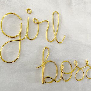May include: Gold wire sculpture of the words "girl boss" on a white background.