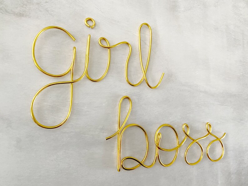 May include: Gold wire sculpture of the words "girl boss" on a white background.