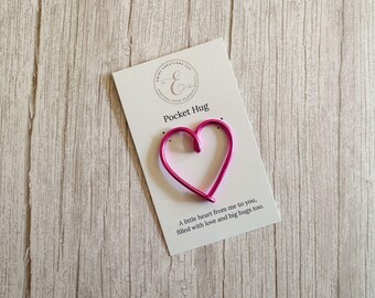 May include: A bright pink wire heart shaped token attached to a white rectangular display card. The card features the text Pocket Hug at the top and the phrase A little heart from me to you, filled with love and big hugs too at the bottom.