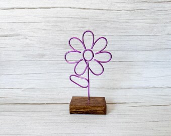 May include: A purple wire sculpture shaped like a flower with eight petals and a single leaf on the stem. The wire flower is mounted into a rectangular, dark brown stained wooden base, providing a stable foundation for the decorative object.