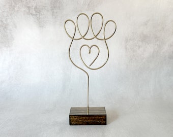 May include: A silver-toned wire sculpture shaped like a paw print with a heart in the center, mounted on a rectangular dark brown wooden base. The wire forms four rounded toe pads at the top and a single heart outline positioned below them.