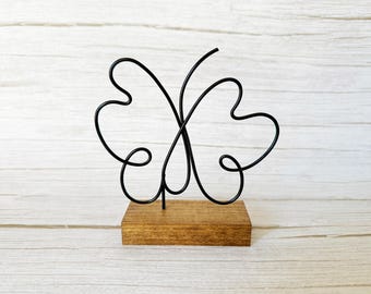 May include: A black metal butterfly sculpture with a minimalist design. The butterfly is mounted on a small, rectangular wooden base. The sculpture is a decorative item, suitable for home decor.
