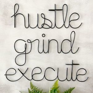 May include: Black metal wire wall art with the words "hustle grind execute" in a cursive font.