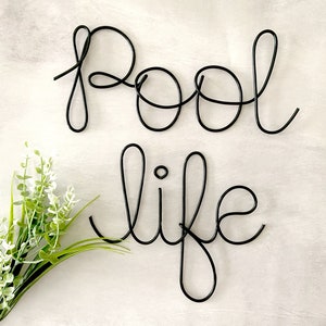 May include: Black wire word art that reads "pool life" on a white background. A small bouquet of white flowers with green stems is in the lower left corner of the image.