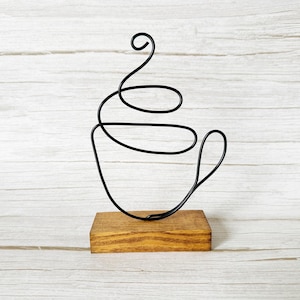 May include: A black wire coffee cup sculpture on a stained wooden base. The cup design is a continuous line, with a handle and steam detail. The sculpture is a decorative item for a kitchen or coffee-themed space.